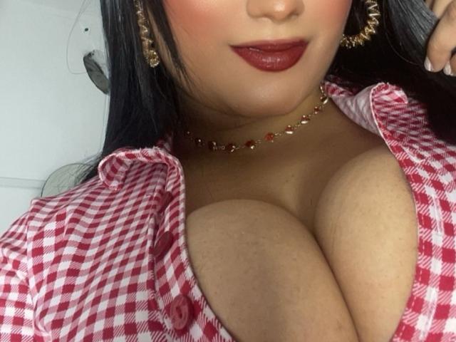 AlexaCooper's large tits are visible in a red and white checkered shirt, with dark red lipstick, gold earrings, and a red bead necklace. Her chest is prominently displayed.