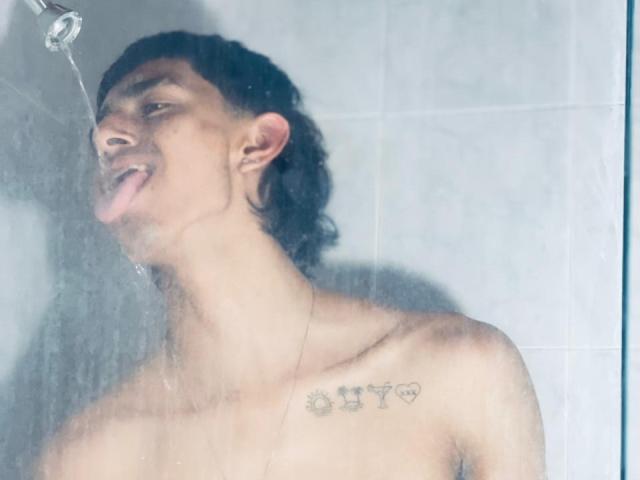 NathanMaxime est sous la douche, de l'eau éclabousse son visage, il a la langue dehors, il est torse nu avec des cheveux noirs bouclés, un petit tatouage sur la poitrine et la peau mouillée, et il regarde l'eau..