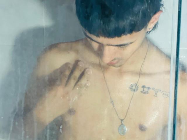 NathanMaxime, zonder shirt, met natte huid, kort zwart haar, naar beneden kijkend, rechterhand op de borst, zilveren ketting met een ronde hanger, tatoeage op de rechter schouder, in een glazen douche..