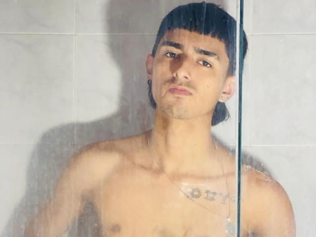 NathanMaxime, shirtless, standing in a shower with water on glass, has dark hair in a straight fringe, brown eyes, small tattoo on chest, and a neutral expression.