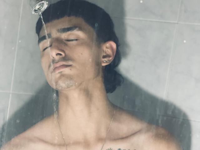 NathanMaxime, zonder shirt, met donker haar in een lage staart, ogen gesloten, water van een douchekop op zijn gezicht, kleine oorbel, licht vochtige huid, neutrale uitdrukking..