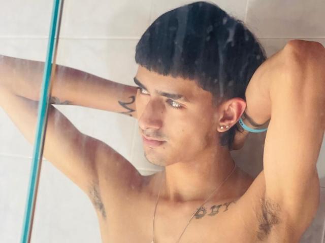 NathanMaxime, met kort zwart haar, armen omhoog, okselhaar zichtbaar, tatoeages op arm en borst, oorbellen en een ketting, met een serieuze uitdrukking, staand onder de douche..