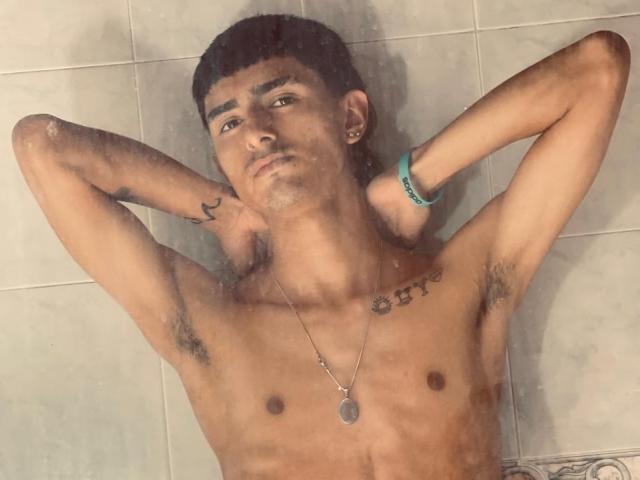 NathanMaxime ist ein Mann ohne Hemd, mit kurzen schwarzen Haaren, den Armen hinter dem Kopf, der Achselbehaarung, Tattoos und einer Halskette zeigt, und steht unter der Dusche..