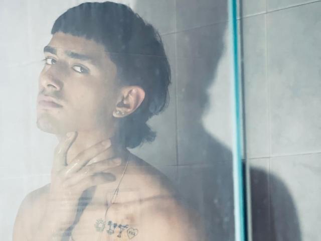 NathanMaxime, sem camisa, com cabelo escuro e ondulado, olha através da porta de vidro do chuveiro, com a mão no pescoço, tatuagem no peito, azulejos cinza claro, sombra visível, com uma expressão ligeiramente irritada..