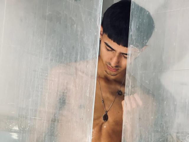 NathanMaxime mit kurzen, schwarzen Haaren, einer Halskette und einem kleinen Brusttattoo, der durch eine nasse, transparente Duschkabinentür späht und mit einem leichten Lächeln nach unten schaut..