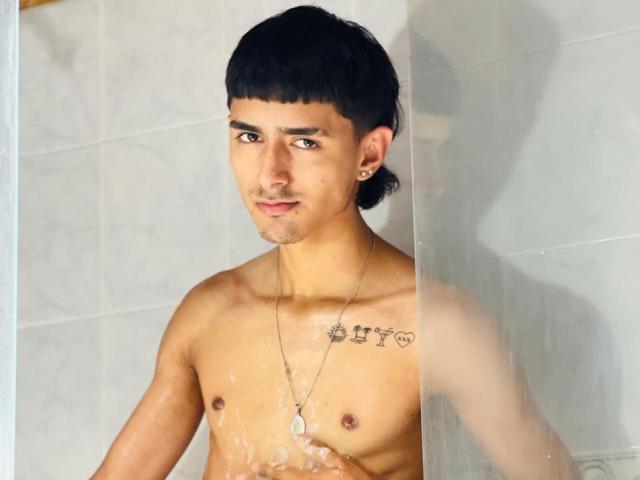NathanMaxime, zonder shirt, met donker haar, een klein borstgedeelte en een tatoeage "HY" op de rechter borstspier, staat onder de douche, met water op zijn borst, en kijkt naar de camera. Hij heeft piercings in zijn oren en draagt een ketting..