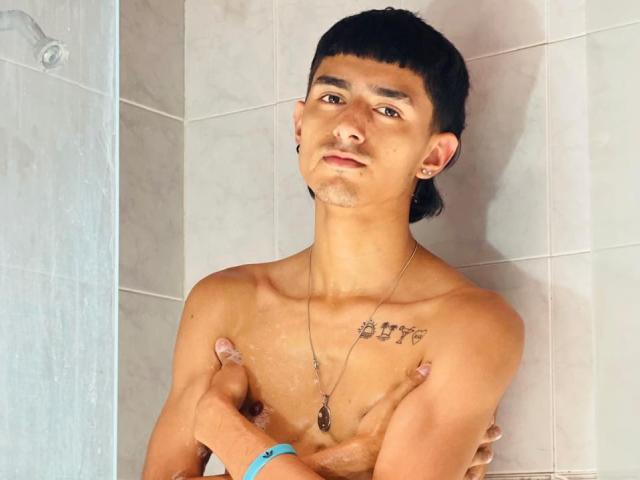NathanMaxime, shirtless, with small tattoos on chest, arms crossed, holding nipples, short black hair, slight facial stubble, necklace, and earring, standing in tiled shower, water droplets.