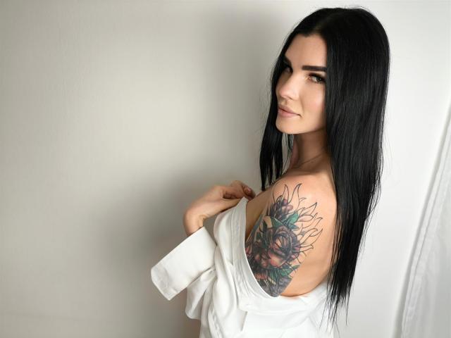 SofiaDream with long black hair, white shirt off-shoulder, showing a sunflower tattoo on her right boob, smiling, looking over her shoulder.