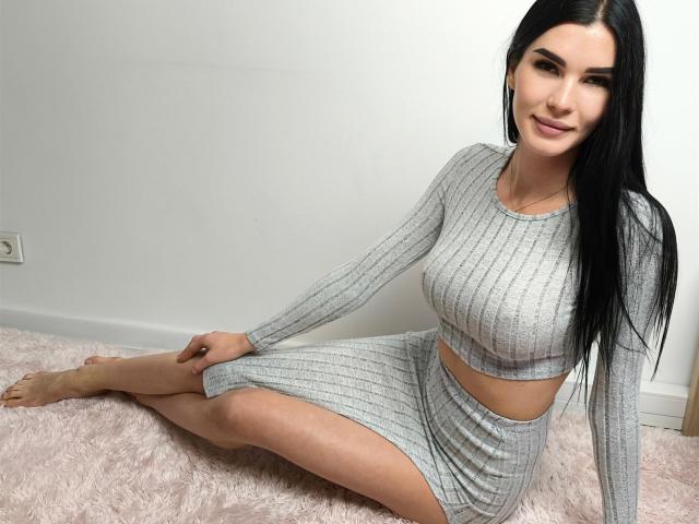 SofiaDream with long black hair, wearing a gray ribbed crop top and skirt, sitting on a pink fluffy rug, showing her tits and ass, smiling at the camera.