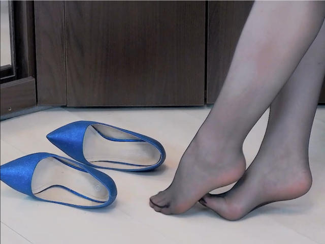 Los pies de MoonBlossom, cubiertos con medias negras transparentes, están descalzos junto a unos zapatos de tacón azul sobre un suelo blanco, con armarios de madera oscura en el fondo..
