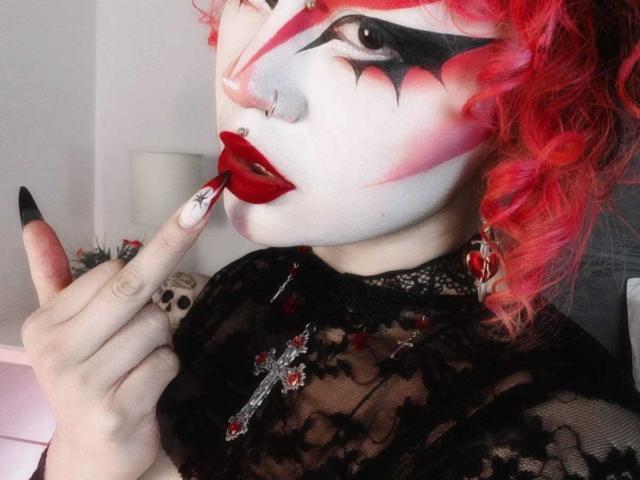 GothVioleth avec des cheveux bouclés roux, un maquillage pâle, un eye-liner noir, du rouge à lèvres rouge, un haut en dentelle noire, un doigt pointé vers la bouche, des ongles rouges et noirs, un anneau au nez et un collier avec une croix..