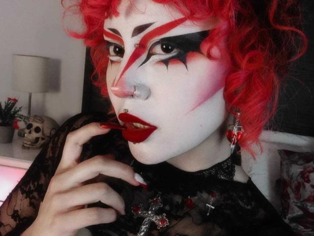GothVioleth tiene el pelo rojo rizado, pintura blanca en la cara con patrones de estrellas rojas y negras, lápiz labial rojo, una blusa de encaje negro, un piercing en la nariz y aretes de cruz, con uñas rojas que tocan la barbilla..