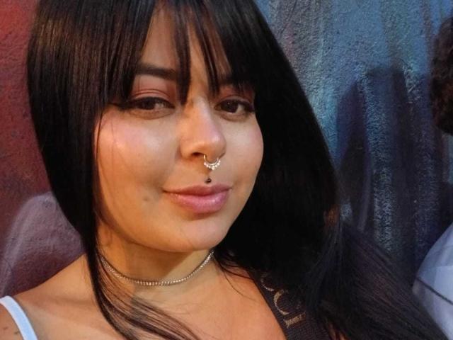 SaraLaJolie ha i capelli neri, un piercing al naso e un piercing al setto con una piccola gemma. Indossa una collana e sorride con un rossetto rosa chiaro..