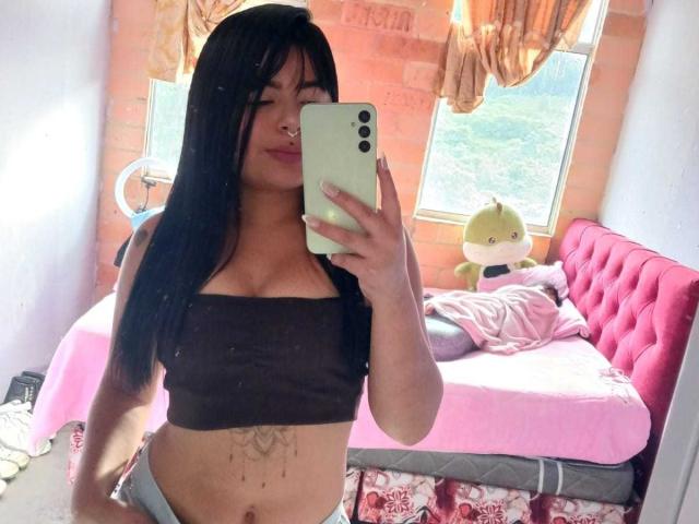 SaraLaJolie, avec ses longs cheveux noirs, son anneau de nez et son haut noir moulant, tenant un téléphone, montrant un tatouage sur le ventre, un lit rose et un jouet vert en peluche, dans une pièce baignée de soleil..