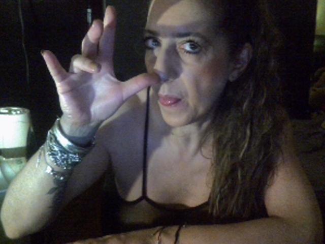 LabelgeCoquine with long brown hair, pink lipstick, and a black spaghetti strap top, making a "rock on" gesture with her right hand, wearing silver bracelets.