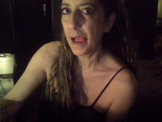 LabelgeCoquine with long brown hair, wearing a black spaghetti strap top, has her mouth open and tongue slightly out, looking surprised, with a can on a dark surface.