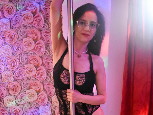 AlanaMoneth with glasses, black lace bodysuit, holding a pole, showing her tits, pink roses background, red curtain, smiling, necklace, right arm raised, medium-length dark hair.