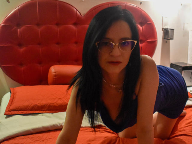 AlanaMoneth with black hair, glasses, and red lipstick, wearing a blue dress, kneeling on a bed with red pillows and a red tufted headboard, looking at the camera.