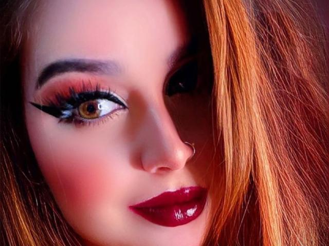 Calen's face, showing a nose ring, red glossy lipstick, and thick black eyeliner on brown eyes with long eyelashes, her orange hair partially covering her right eye.