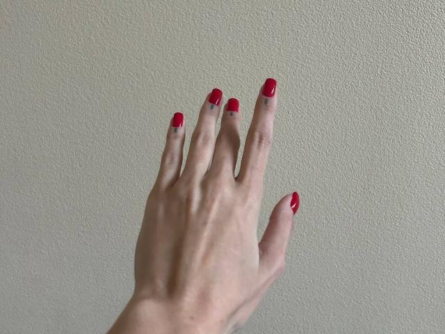 La mano di MoonLiza, con le unghie dipinte di rosso, le dita leggermente divaricate, appoggiata contro un muro spoglio. La sua pelle è pallida e le sue unghie sono curate..