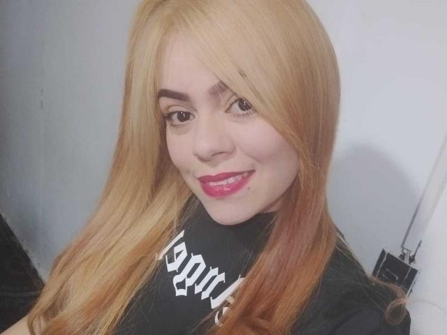 KataBlonde has blonde hair, pink lipstick, and a cute smile. She's wearing a black shirt with white text. She has a small beauty mark on her right cheek.