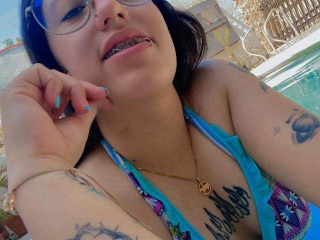 NatalieHarris with glasses, black hair, blue nails, metal lip piercing, tattoos, gold necklace, blue patterned bikini, hand near mouth, pool in background, sunny day.