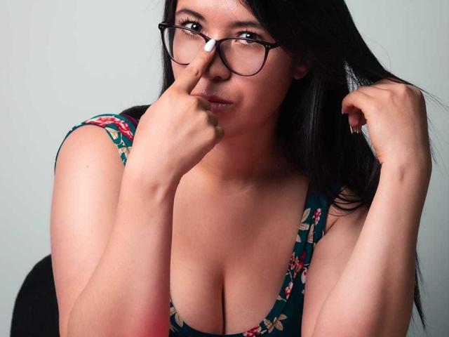 LizBellaa with glasses, black hair, and floral dress, touching nose with finger, showing cleavage, looking slightly annoyed, holding hair with other hand.