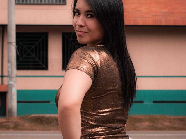 LizBellaa with long black hair, looking over her shoulder, wearing a shiny brown tight-fitting top, smiling slightly, her butt is partially visible, standing outside.