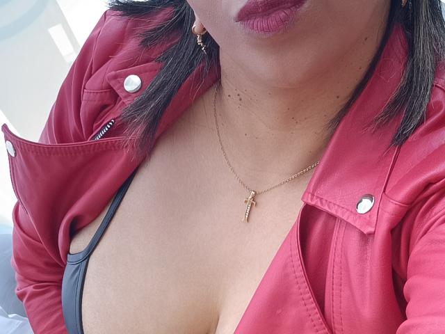 La veste en cuir rouge de Madame Victoria est ouverte, laissant apparaître son soutien-gorge noir et sa poitrine. Elle porte un rouge à lèvres foncé, un collier avec une croix en or, et sa peau est de couleur marron clair..