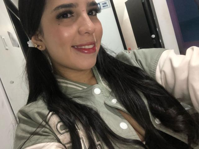 AlanaReyes has long black hair, green varsity jacket, red lipstick, and a cute small mole on her cheek, smiling at the camera, showing her tits slightly through the jacket.