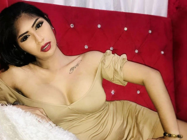 Sensualbunny-hot with black hair, red lipstick, and tan dress, showing off her tits and tattoo on her chest, lying on a red tufted couch with white pillows.