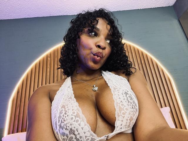 LisaMendez with curly black hair, wearing a white lace halter top, showing large boobs, pouty expression, and a silver necklace, against a wooden headboard.