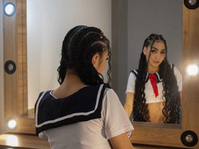 LylaPeach with curly black hair in a Japanese schoolgirl uniform, facing a mirror, reflecting her face, small tits, and a red necktie, in a lit wooden-framed mirror.