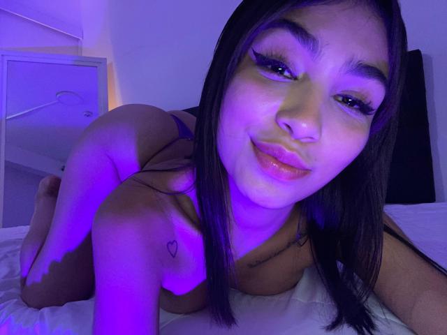 LylaPeach is on all fours, naked, with purple lighting. She has long dark hair, thick eyelashes, and a heart tattoo on her left shoulder, showing off her ass.
