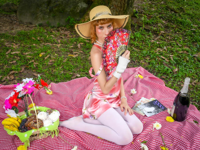 AuraFire with red floral hat, pink dress, white tights, holding fan, sitting on red checkered blanket, surrounded by picnic items, bottle, flowers, and book, in a forest.