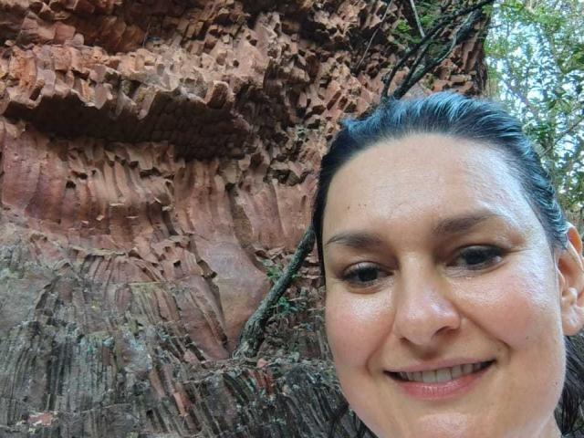 LadySelene's face with a smile, brown eyes, and black hair tied back, standing in front of a rugged, reddish-brown rock wall with visible textures and small plants.