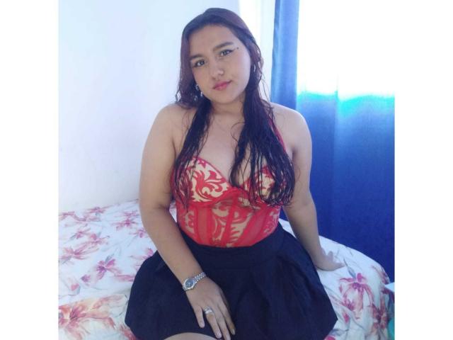 ElsaSantos with wet brown hair, wearing a red lace top showing her boobs and a black skirt, sitting on a bed with a floral bedsheet, blue curtain behind.