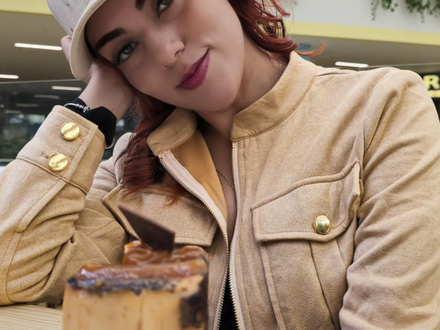 ZoeeMar with red hair, beige jacket, gold buttons, white cap, dark lipstick, smiling at chocolate cake with chocolate stick, hand on cheek, black wristband, indoors.