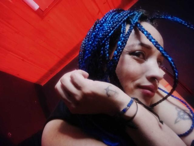 DalilaMusa with blue braids, nose ring, and tattoos, smiling, showing her tits, arm raised, black top, red ceiling, and a bracelet, in a dimly lit room.
