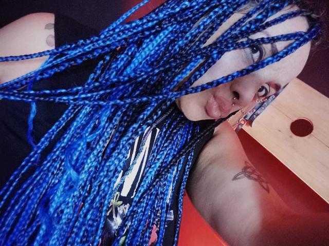 Dalila Musa, con trenzas azules brillantes, sacando la lengua, vistiendo una camisa negra, con un tatuaje visible en el brazo, un piercing en la nariz, y mirando a la cámara..