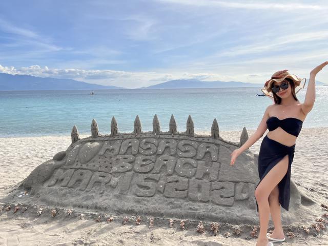 StacyMalena with boobs in a black strapless top and skirt, standing by a sandcastle with a spiked top, wearing sunglasses, and a hat on a beach.