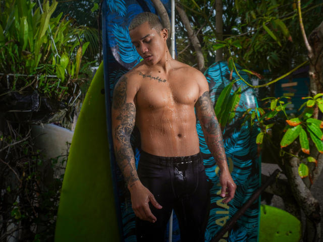 CristianoGabi, shirtless with wet skin, tattoos on arms and chest, black shorts, standing by a blue and green surfboard, surrounded by greenery, looking down with a slight smile.