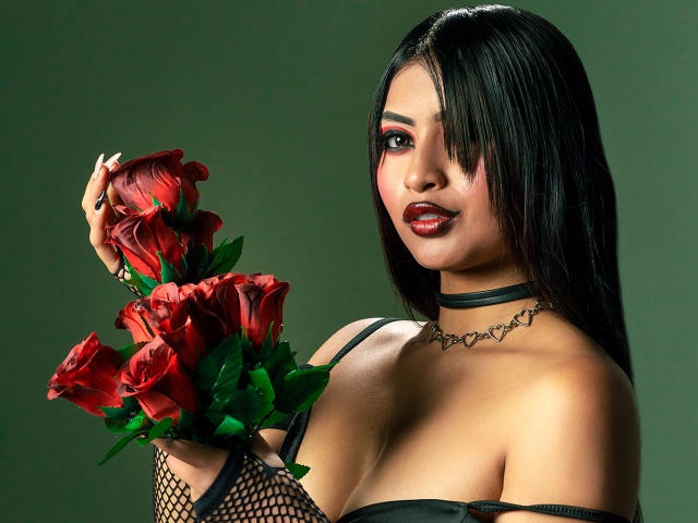 EmmaaRodriguez with black hair, red lipstick, fishnet arm, black choker, holding red roses, showing her big tits in a black strap top. Green background.
