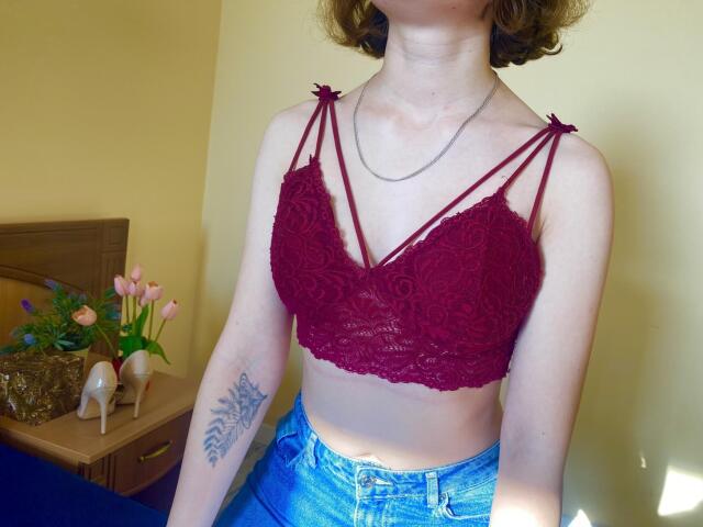 Steisii with pale skin, short curly brown hair, wearing a red lace bralette with strappy details, blue jeans, and a necklace, showing a tattoo on her right arm.