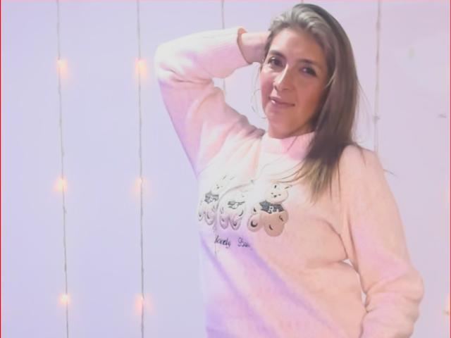MichelleDumont with brown hair, smiling, wearing a pink sweater with cute cat faces, hand in hair, standing, soft pink and white lighting, warm fairy lights in background.