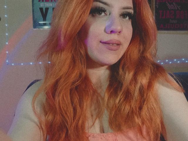 SpicyGrace ha lunghi capelli rossi ondulati, labbra piene e indossa un trucco per gli occhi molto marcato. Sta guardando la telecamera con un leggero sorriso, mostrando il seno..