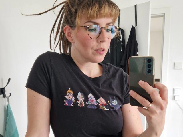 GypsyPageXXX - une femme à la peau bronzée, avec des dreadlocks bruns et gonflés, des lunettes et un t-shirt noir avec des personnages de dessins animés, prenant un selfie devant un miroir avec un téléphone vert, montrant ses seins..