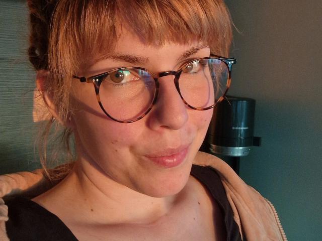 GypsyPageXXX-hot with puffy lips, tortoiseshell glasses, light brown hair in a messy bun, and a smug expression, wearing a beige zip-up and black top.