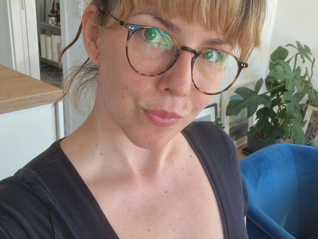 GypsyPageXXX-hot with tortoiseshell glasses, green-eyed reflection, light freckles, pouty lips, blonde bangs, black V-neck, slight smile, blue chair in background.