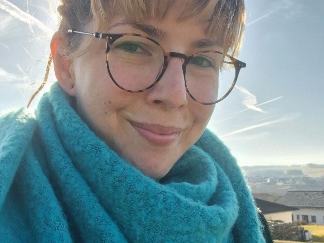 GypsyPageXXX-hot with tortoiseshell glasses, blue fuzzy scarf, and light brown hair, smiling at the camera, sunlight in background, clear blue sky with white contrails.